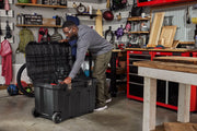 A person is standing in a workshop or garage, using a large black rolling storage chest with an open lid. The workspace is organized with shelves, tools, tool chests, and various items like backpacks, wood pieces, and other equipment visible in the background.